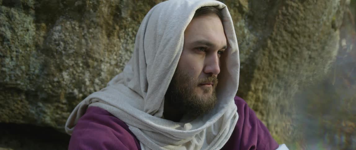 A performer is dressed as jesus and sitting next to a rock