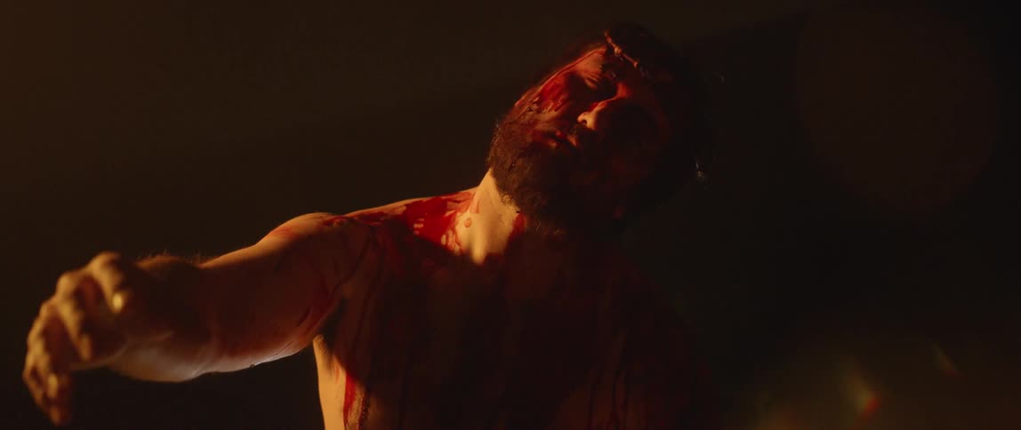 A shirtless man covered in blood reaches his arm and hand out