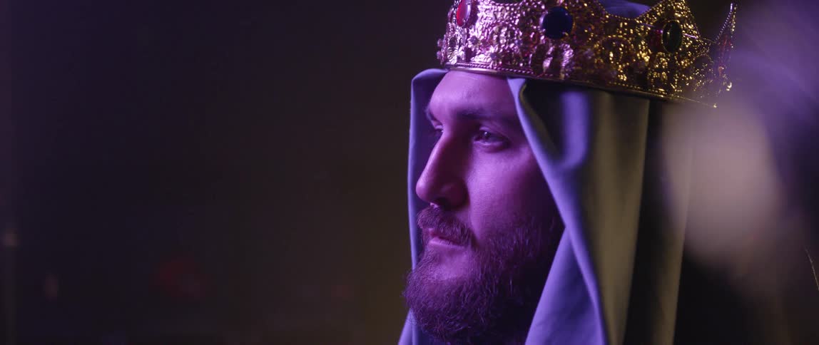 Man wearing crown and head covering stands staring