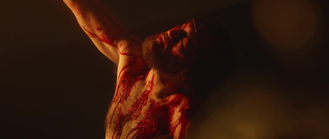 A bearded man stands in the darkness while covered in blood and with a swollen eye