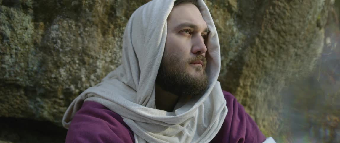 A bearded man has his head covered in a cloak as he sits and looks contemplative