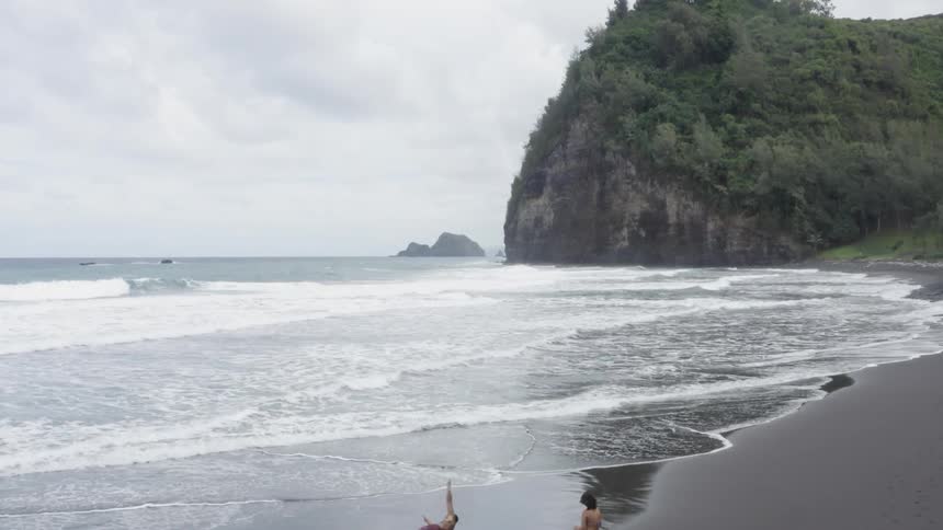Man and woman lie down on the beach and man rolls around next to the ocean on a cloudy day