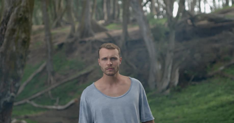 Young blonde man stands and looks at the camera in the forest