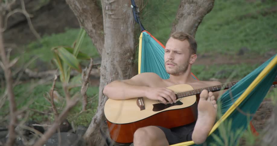 Shirtless male laying in a hammock strums on a guitar as he looks around