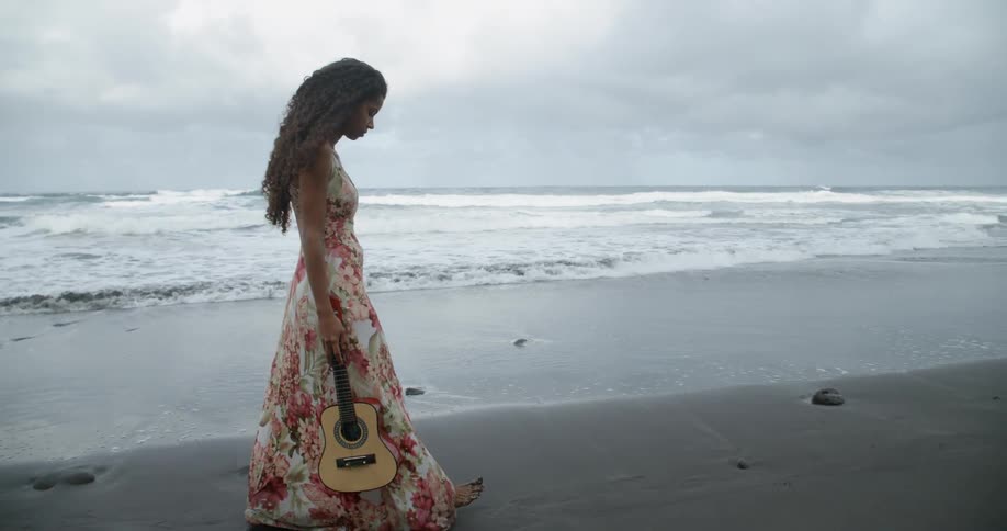 Black woman holding a guitar walks on the wet sand beach next to the sea