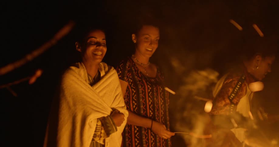 Two women gathered around a bonfire cooking hot dogs at night