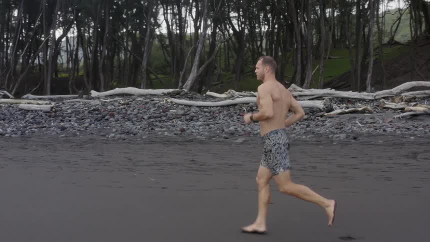 Shirtless and barefoot man runs along the a beach covered in dark sand