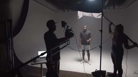 A man stands alone on a soundstage and talks while be filmed and recorded