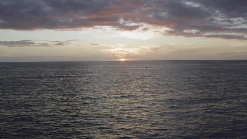 In the sky above the wide open ocean the sun is barely above the horizon under a cloudy sky