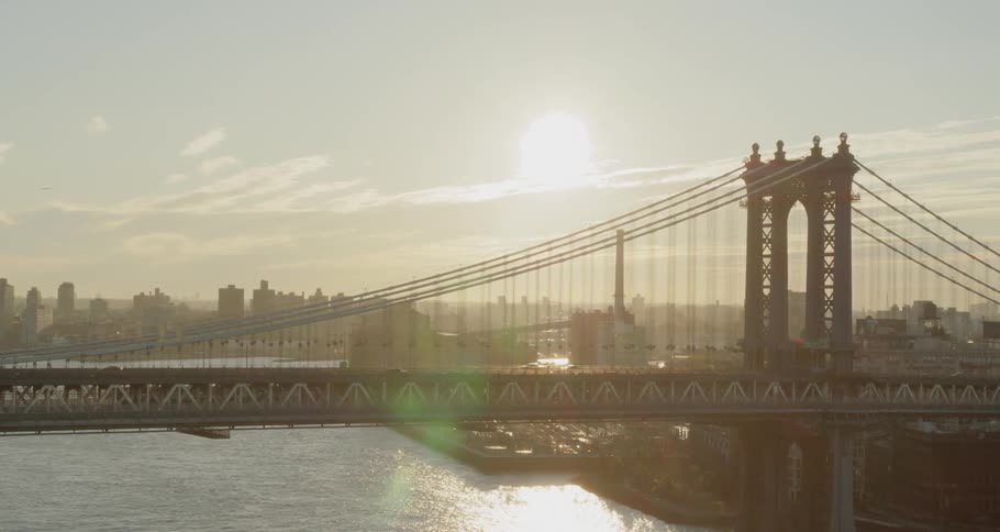 The sun shines behind a suspension bridge that spans a river between two metropolitan areas