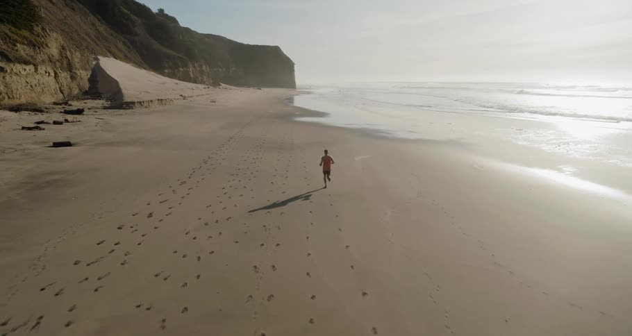 Man jogs along the sandy beach next to the ocean while the sun shines on the sand
