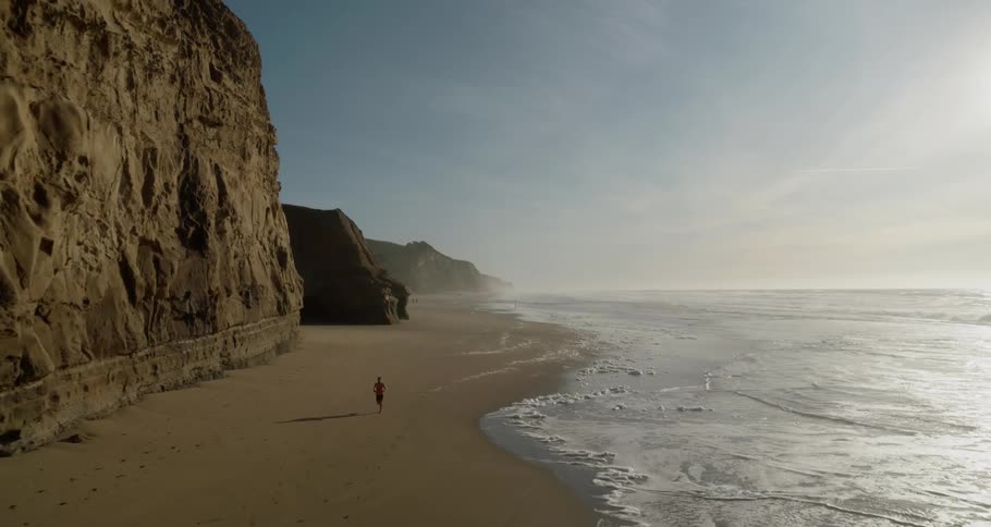 A person jogs beside an ocean coast with tall mountains along the beach