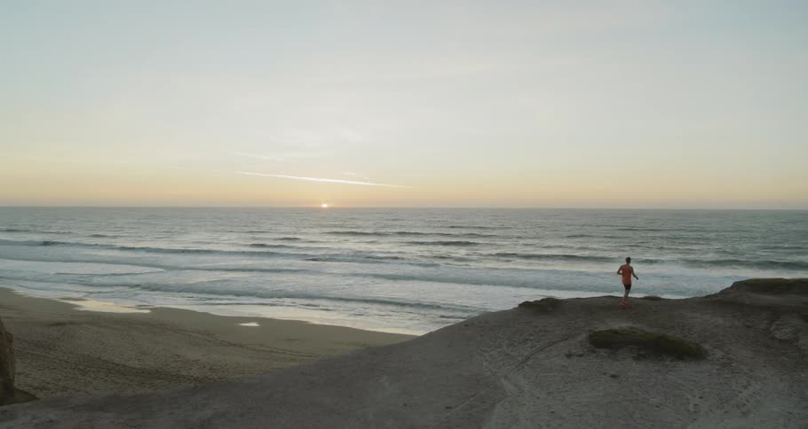 Man runs up to the cliffside and looks at the vast ocean at sunset