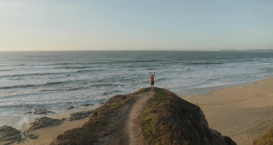 Man runs to the cliffside and raises arms in victorious gesture with the view of coast and ocean