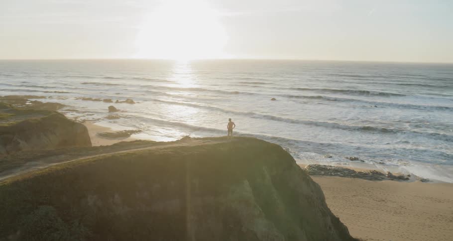 Man stands on top of a hill overlooking the ocean under a sunny sky