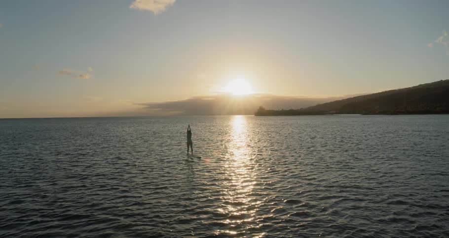 A woman stands atop a long surfboard while floating on the ocean as she stretches her arms above her head