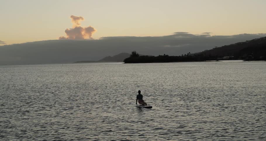 A woman stretches and holds a pose on a surfboard in the water at sunrise