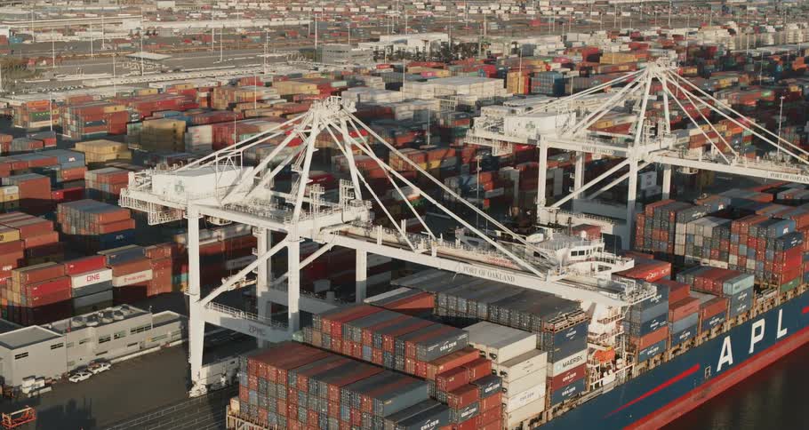 A large freight shipping vessel is docked at a shipping yard under two massive cranes used for moving freight containers