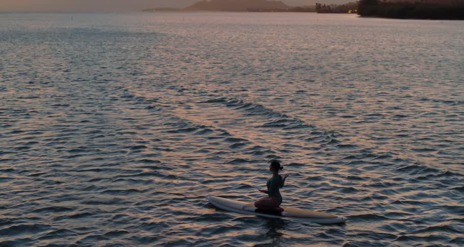 While floating on the ocean water a woman kneels atop a long surfboard and holds her hands outward in a yoga pose