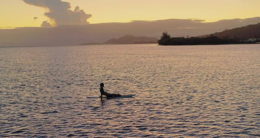 A woman stretches and holds a yoga pose on a surfboard in the water at sunset
