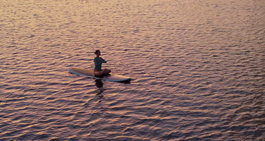 A woman sits on a surfboard in the water meditating at sunset