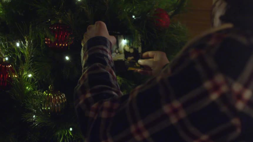 A woman in a plaid shirt approaches a christmas tree and places a photo between the branches