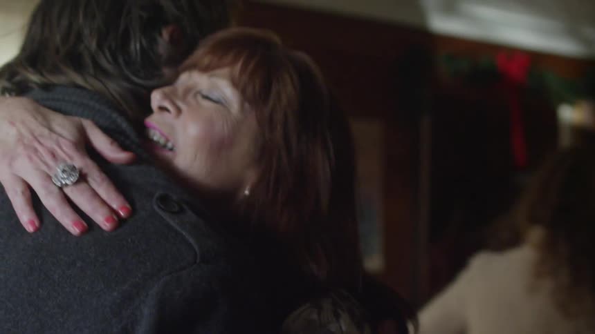 An older woman greets and welcomes a large family inside with smiles and hugs