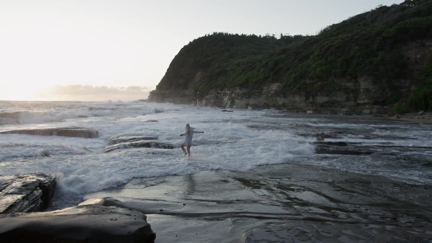A woman spins in a circle on a rocky beach