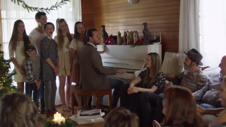 A large family gathering at christmas with some seated while others are standing and singing along with the music being played on the piano