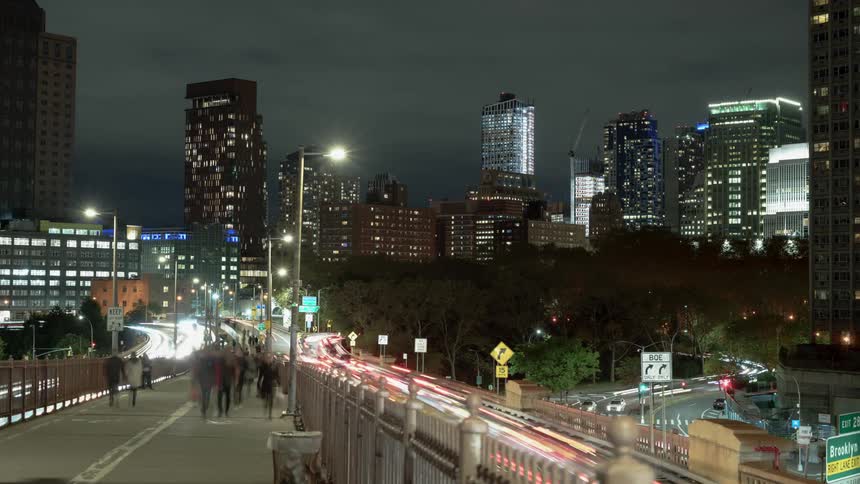 People walk on a pedestrian bridge above traffic moving to and from a city at night