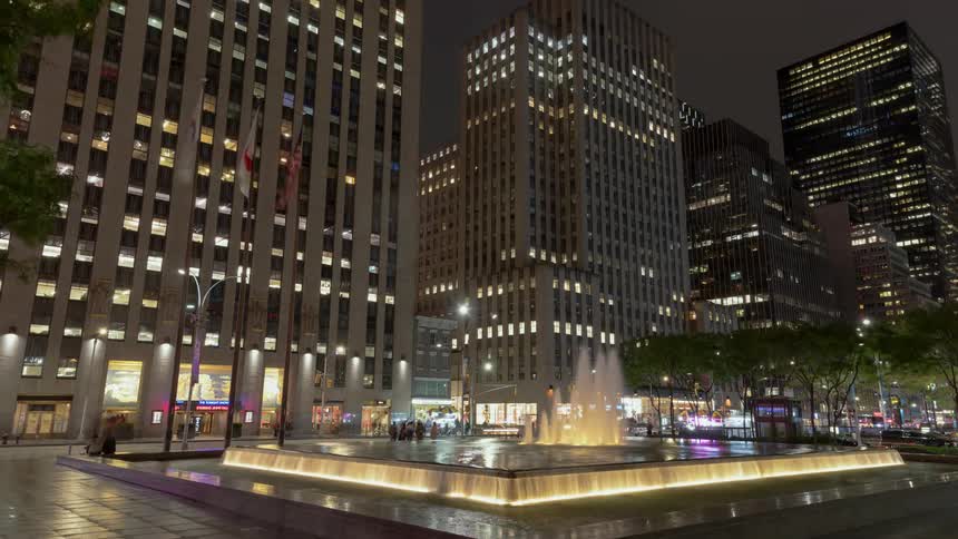 People and traffic move around a large fountain the the downtown of a city