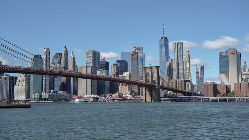 A bridge over a river with boats passing by with the city skyline in the background