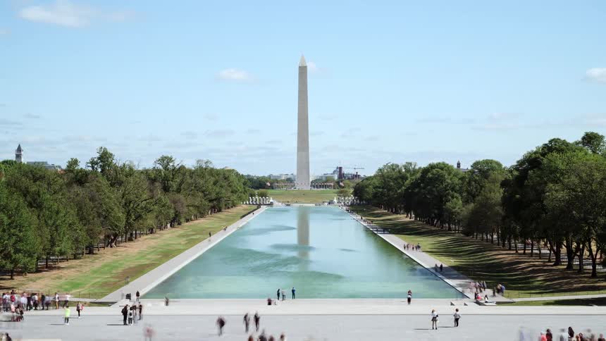 People walk around a lake near a tall monument by the trees
