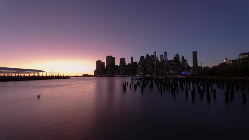 A city skyline is reflected in the water as it turns from day to night