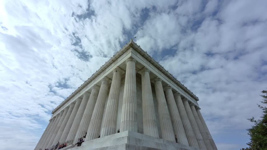 People sitting standing and walking on a monument with pillars with the sky and clouds above