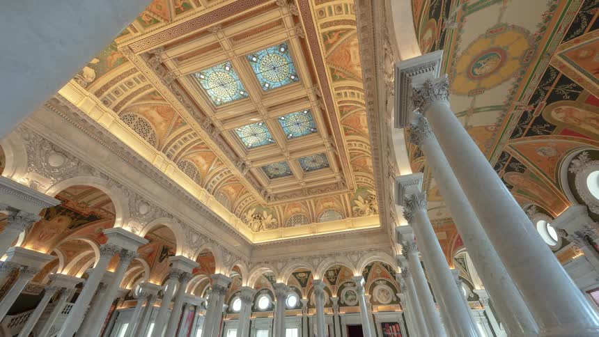 The stained glass ceiling and columns of the library of congress as people walk up and down the stairs