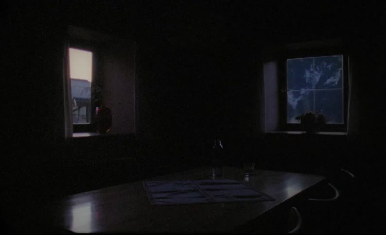 A dining room table in a dark room with someone walking outside seen through the windows