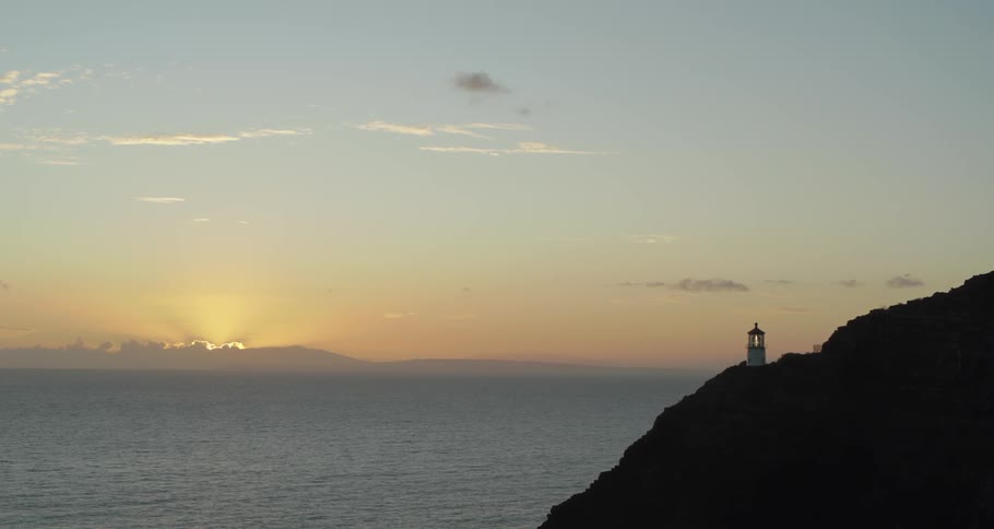 A light house sits on a hill overlooking the sun setting over the water