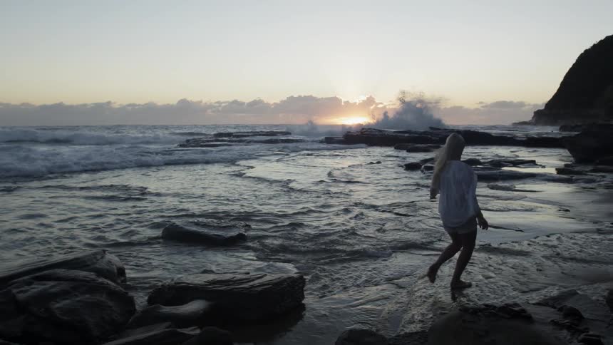 A woman hopping between large rocks on the shoreline during sunset
