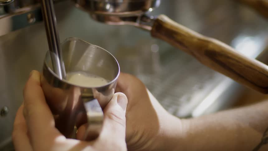 A person holds a small pitcher and steams milk at an espresso machine
