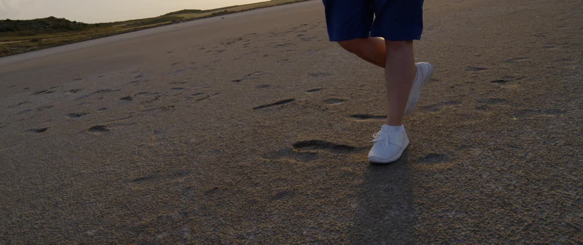 A man wearing shorts and sneakers leaves footprints as he walks on hard packed sand at sunset