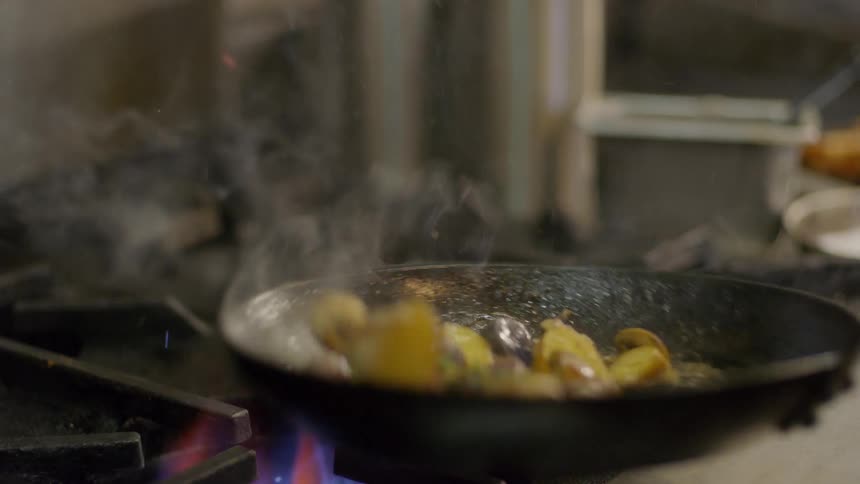 A person shakes a frying pan of food over a flame on a gas stove