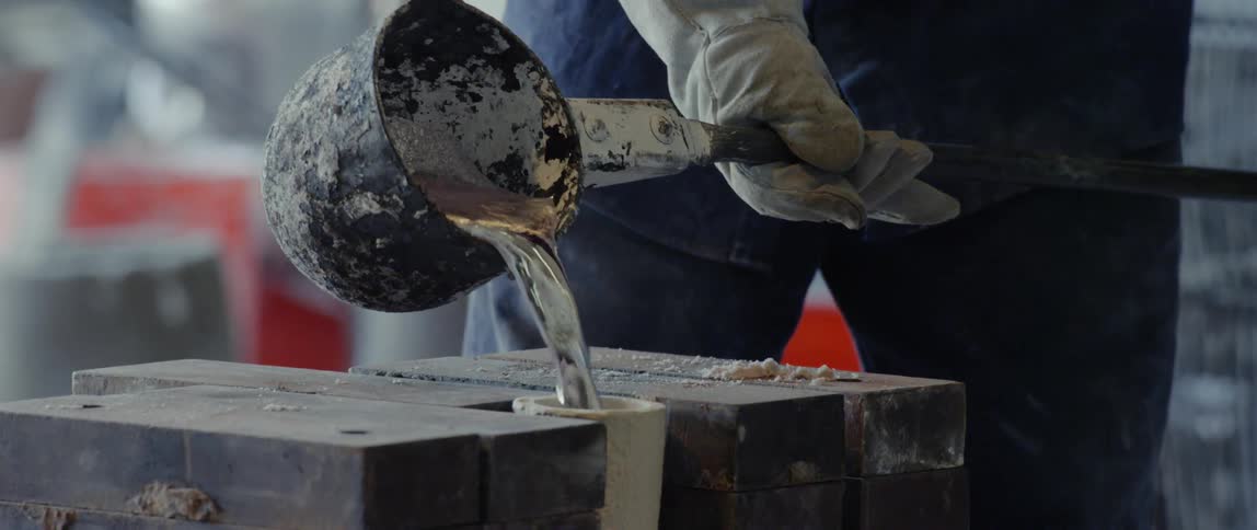 A worker pours molten metal from a ladle into a form
