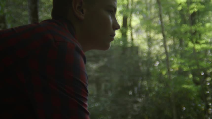 A boy sits leaning forward with elbows resting on his knees looking out at the surrounding trees