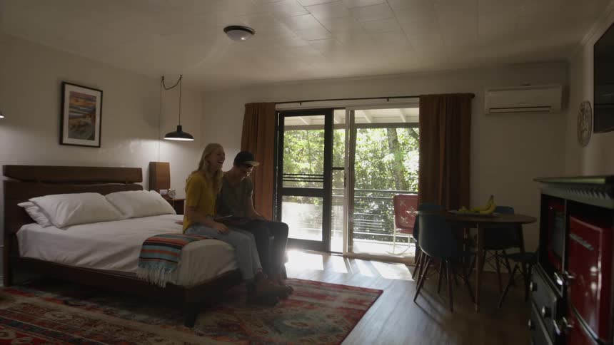 A man and woman sit on the end of a bed and laugh as they look at a book together