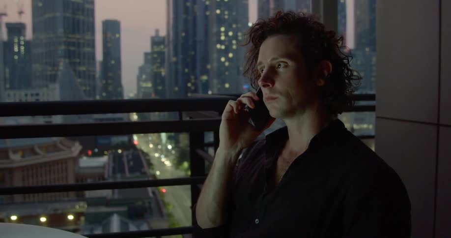 A man talks on his phone and grimaces while on a high rise balcony overlooking the city at dusk