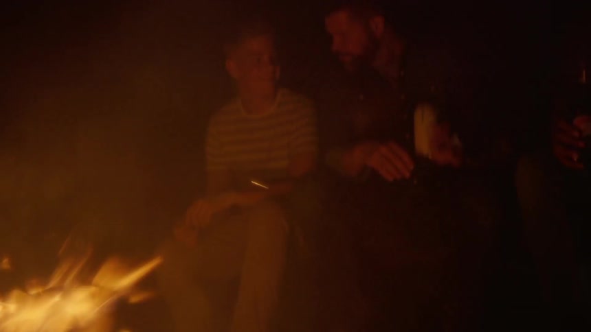 Several people sit around an open fire while one is roasting a marshmallow on a stick