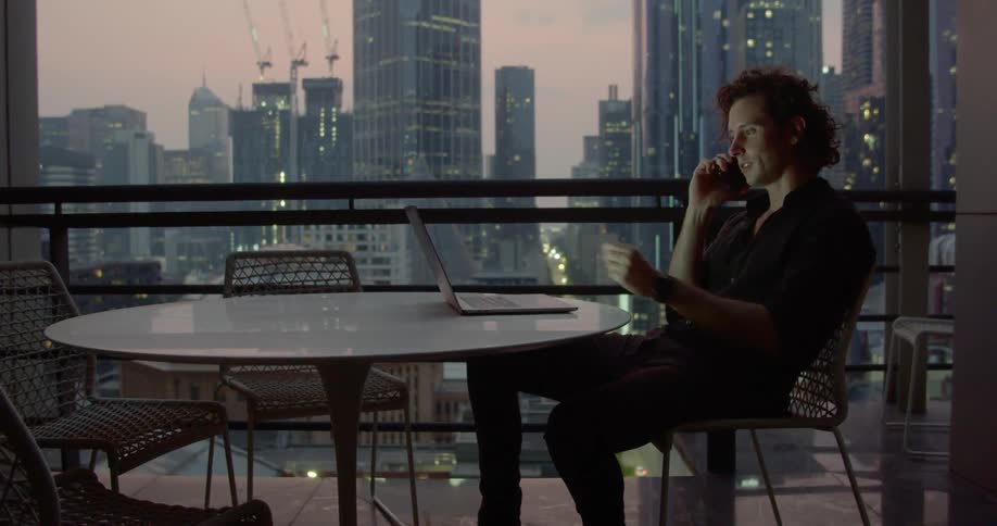 A man sits at a table with a city skyline behind talking on a phone and scrolling on a laptop