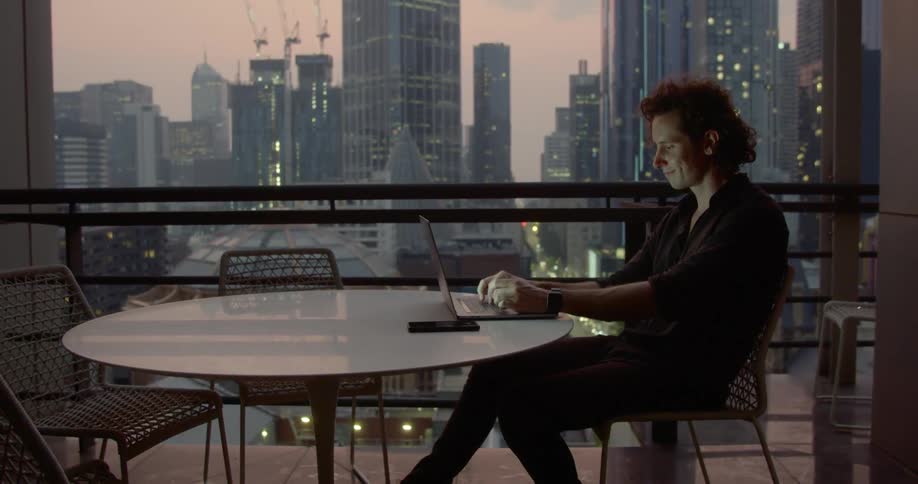 Man works on laptop at table next to wide window with view of city