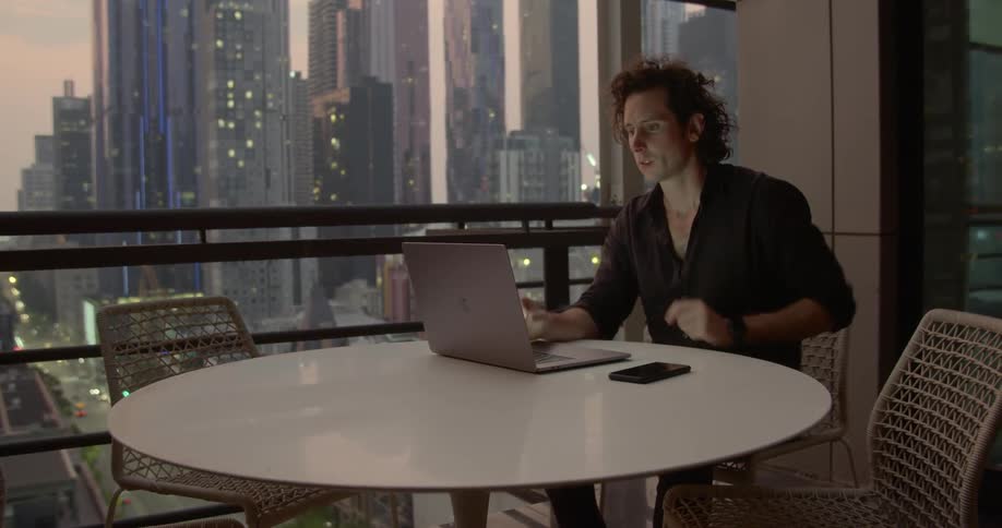 A curly haired man sit at a table next to a window with a view of countless sky scrapers